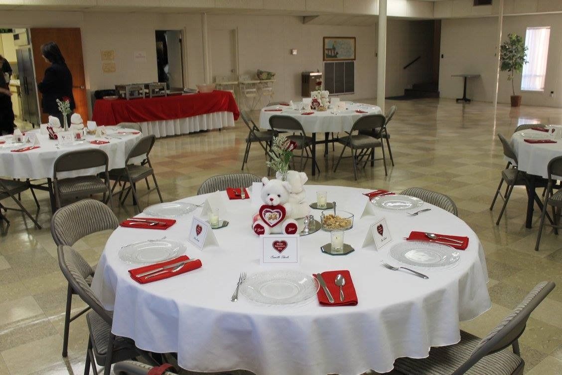 Round banquet tables set for an event in a bright hall, with white tablecloths and red napkins.