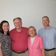 Four people standing together indoors, smiling in front of a plain wall.
