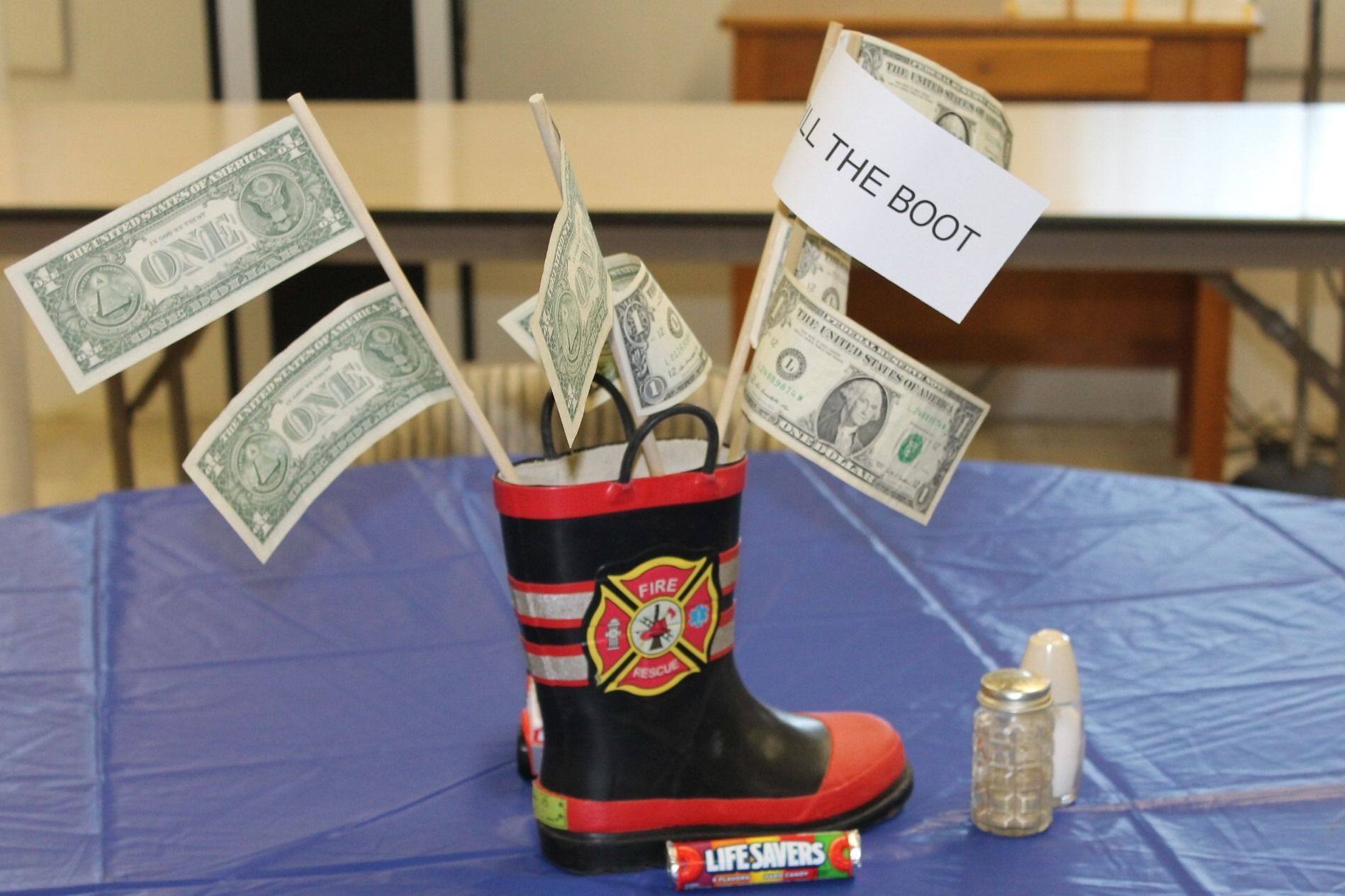A firefighter boot-shaped tip jar with dollar bills and coins on a blue table
