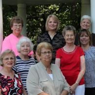 Seven women posing together outdoors, smiling in front of a building.