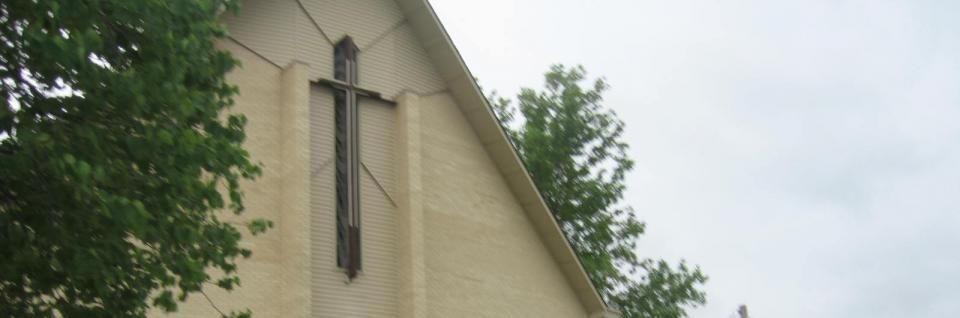 Tall beige church wall with a cross and a stained-glass window against trees and cloudy sky