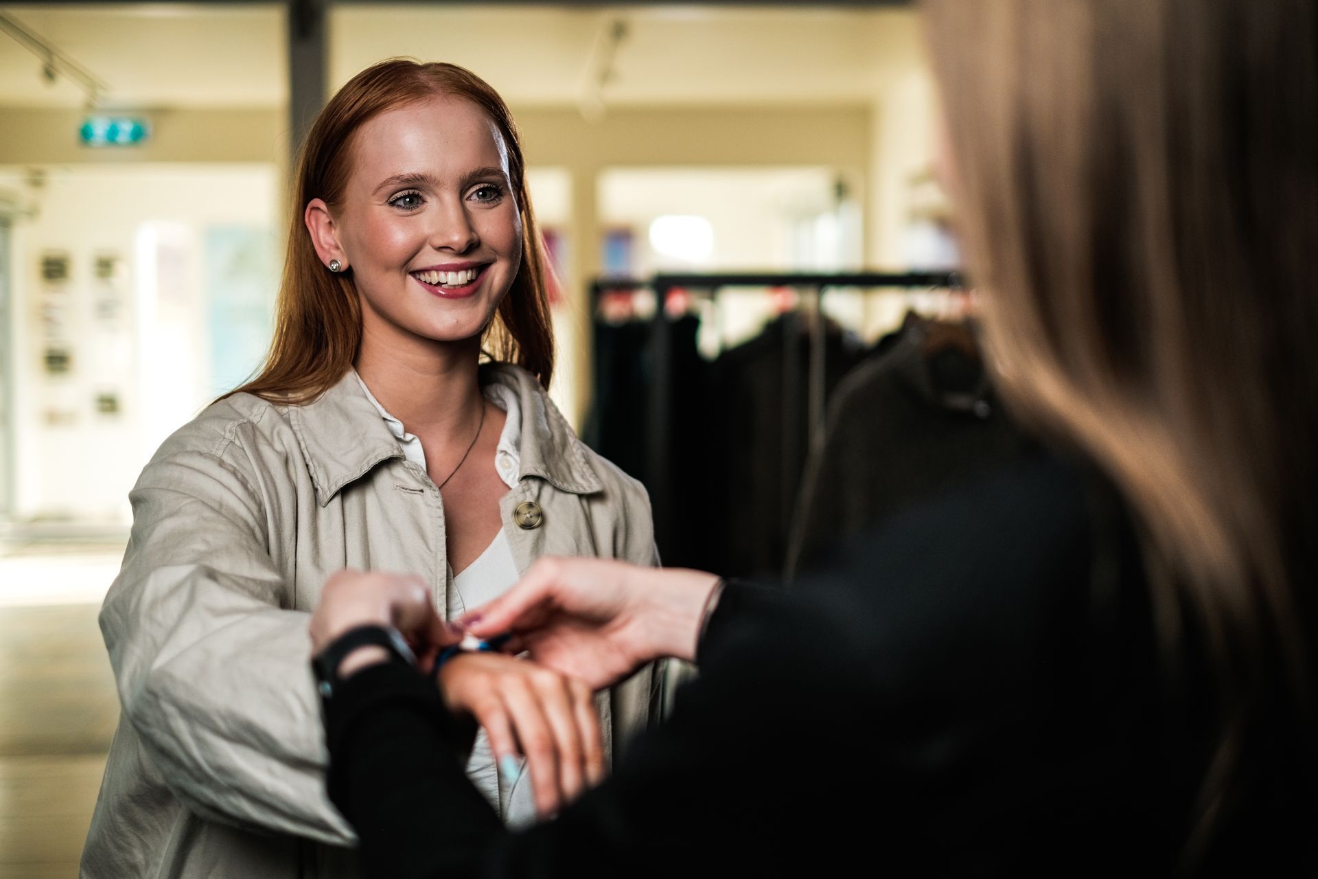 Woman with red hair smiles as another person adjusts a watch on her wrist, in a store setting.