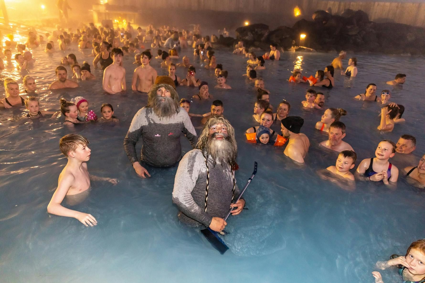 People relax in a blue thermal pool, some with long beards, at night, with fog rising from the water.