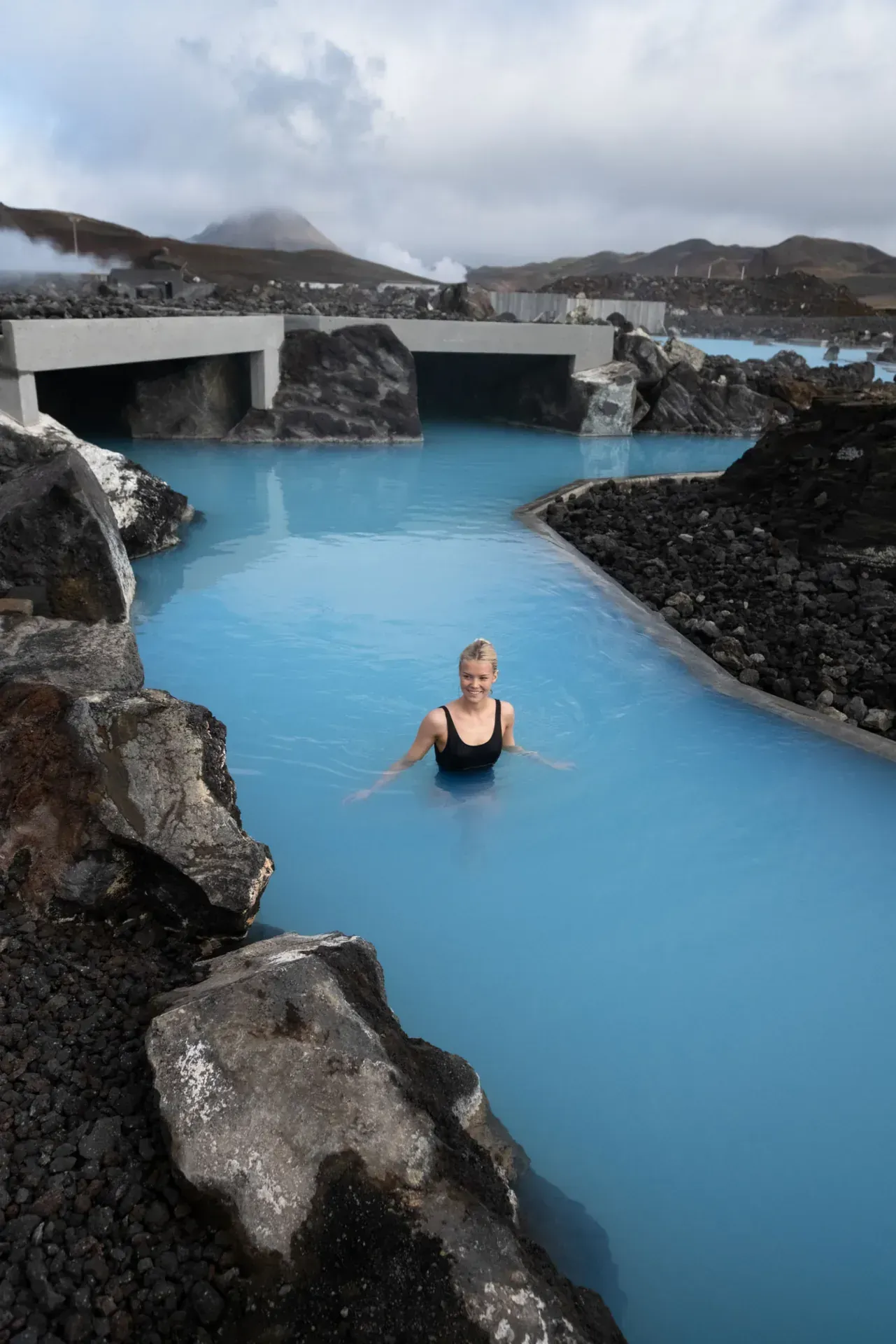 Aerial view of the Blue Lagoon in Iceland: turquoise water, black lava rock, steam rising, cloudy sky.