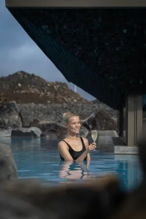 Woman in pool reaching for drink, raising a toast. Light blue water, cabin window.
