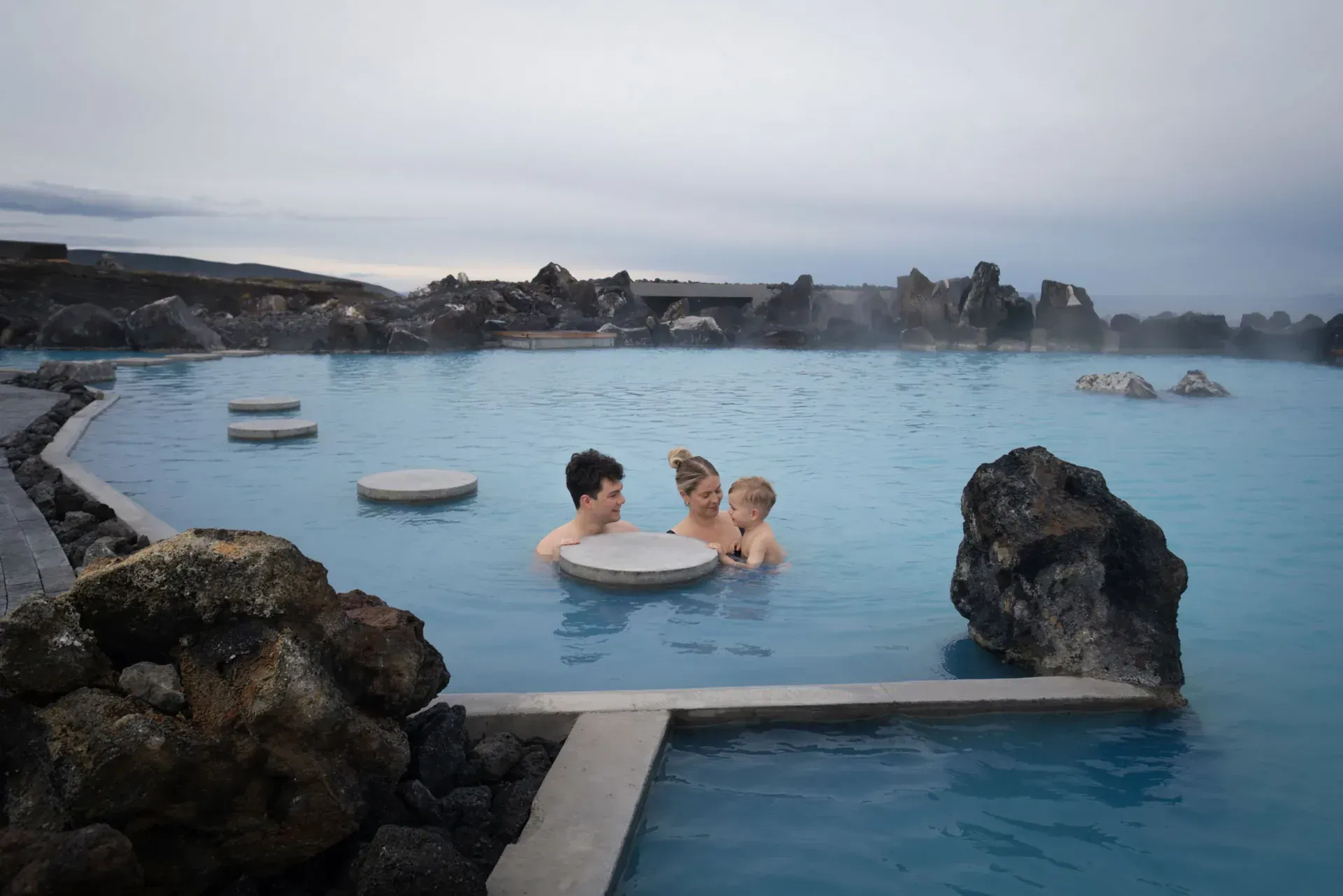 Aerial view of the Blue Lagoon in Iceland: turquoise water, black lava rock, steam rising, cloudy sky.