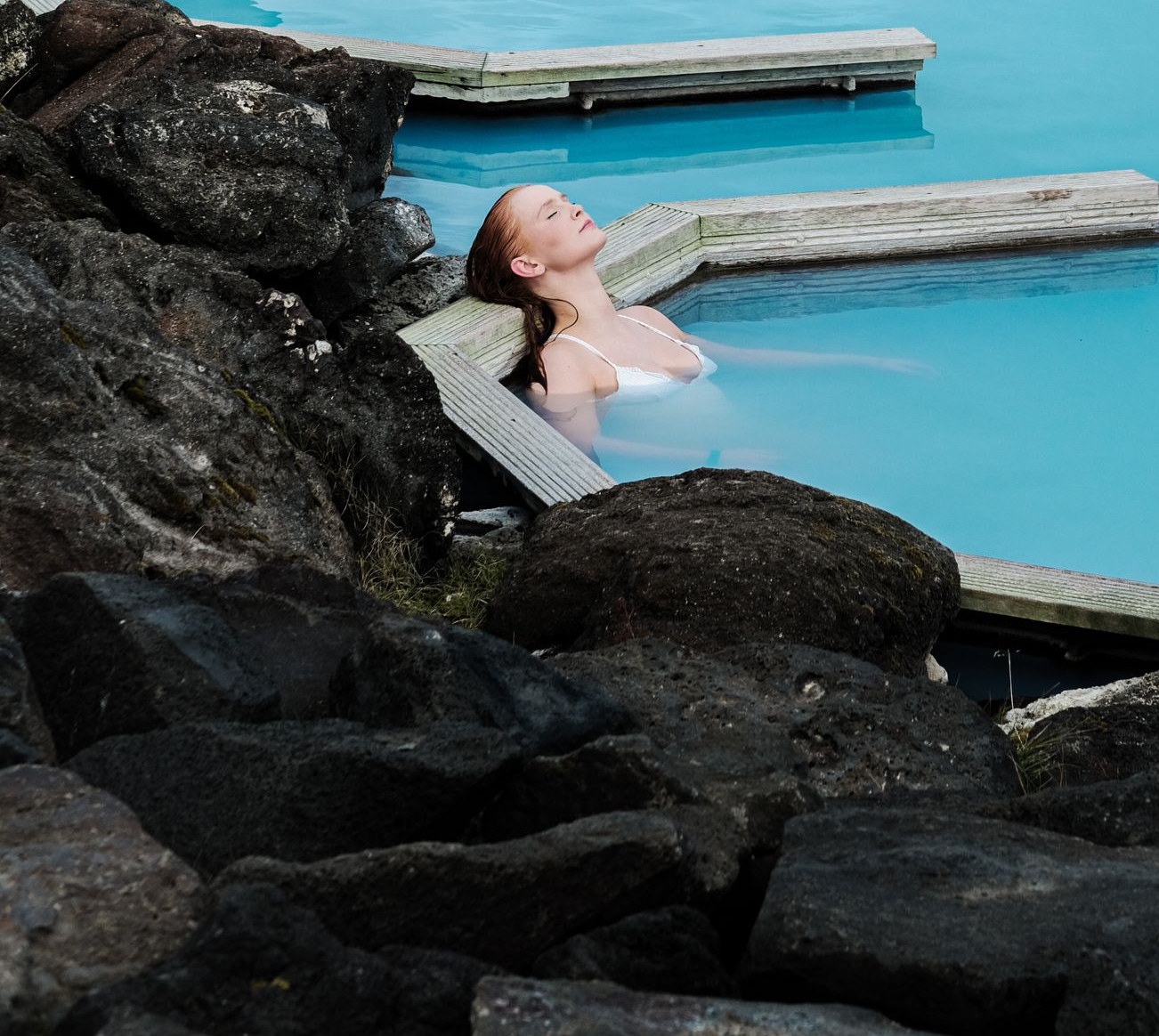 Kona slakar á í bláum jarðhitabaðspotti, hallar sér á brún, með lokuð augu. Umkringd dökkum klettum.