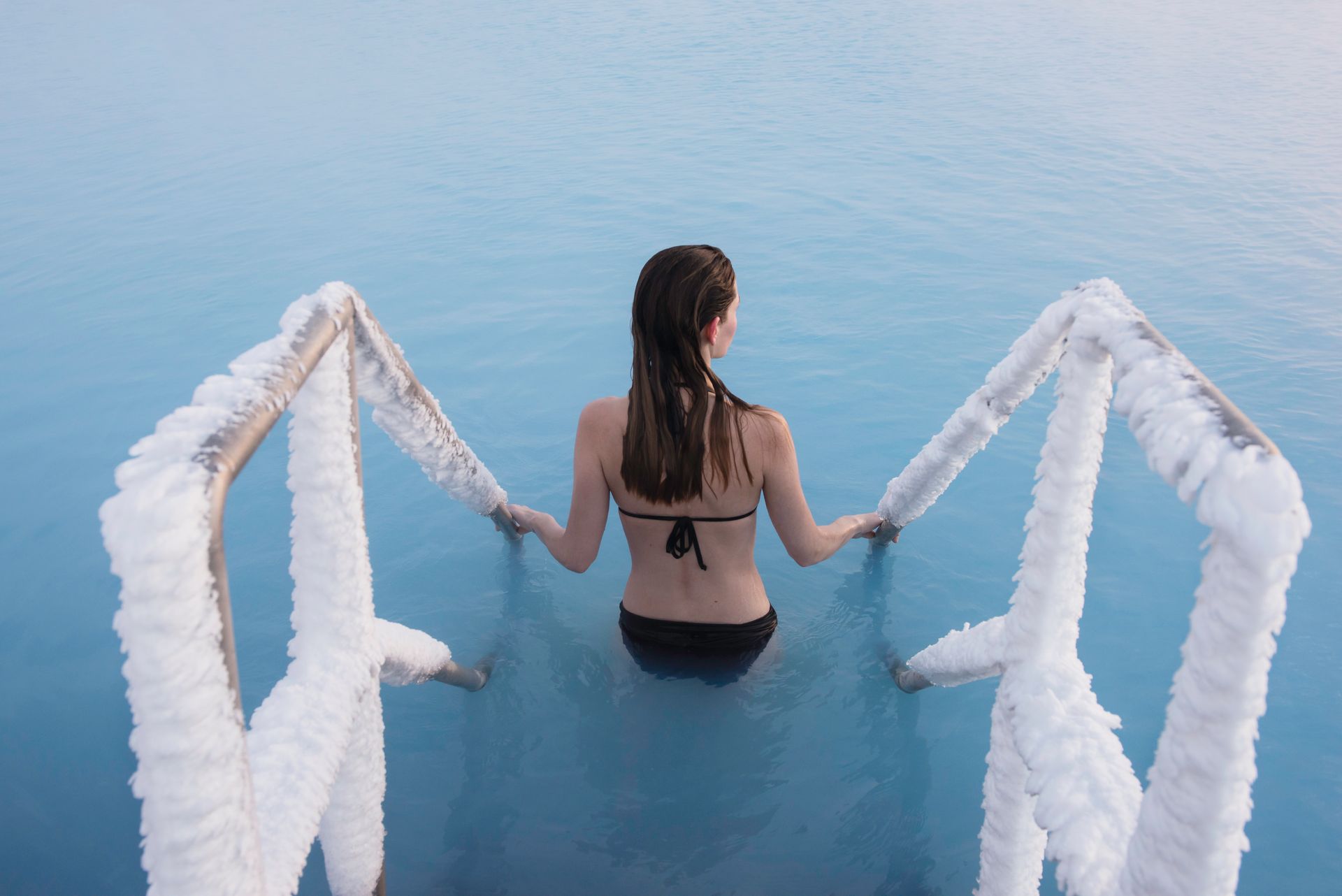 Woman in a black swimsuit, entering blue thermal waters from ice-covered stairs.