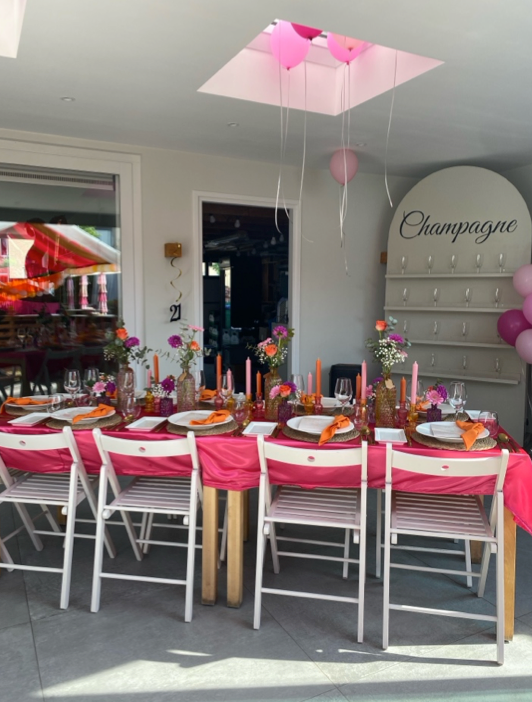 Een feesttafel versierd in roze en wit met ballonnen en een hangende roze paraplu als middelpunt.