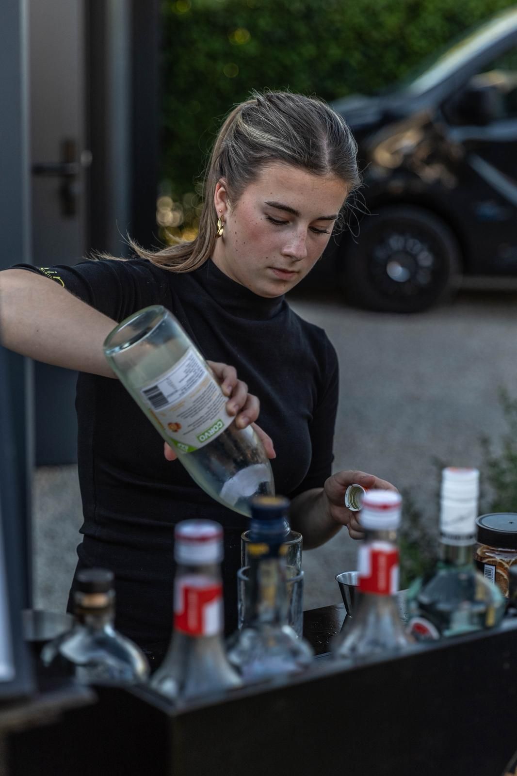 Een barman schenkt sterke drank in een cocktailshaker bij een buitenbar, met flessen in de buurt.