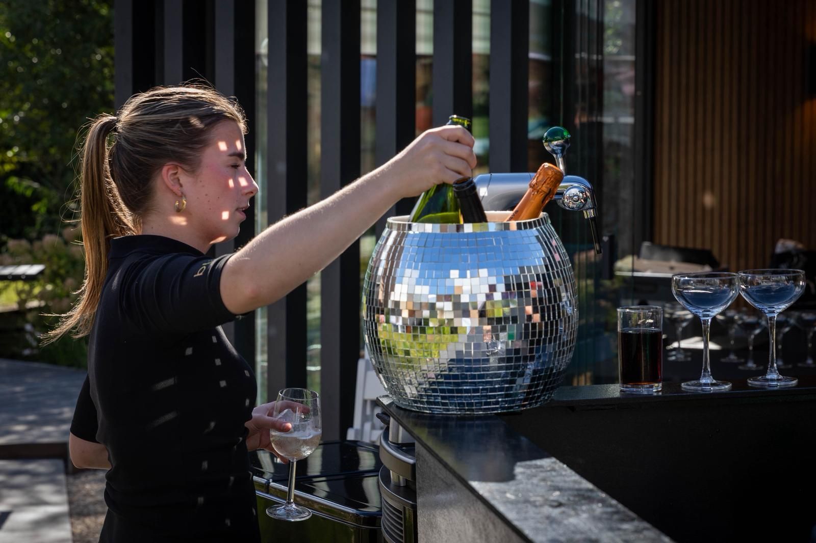 Een barman schenkt een drankje in een ijsemmer met spiegelglas buiten, naast glazen op een bar.
