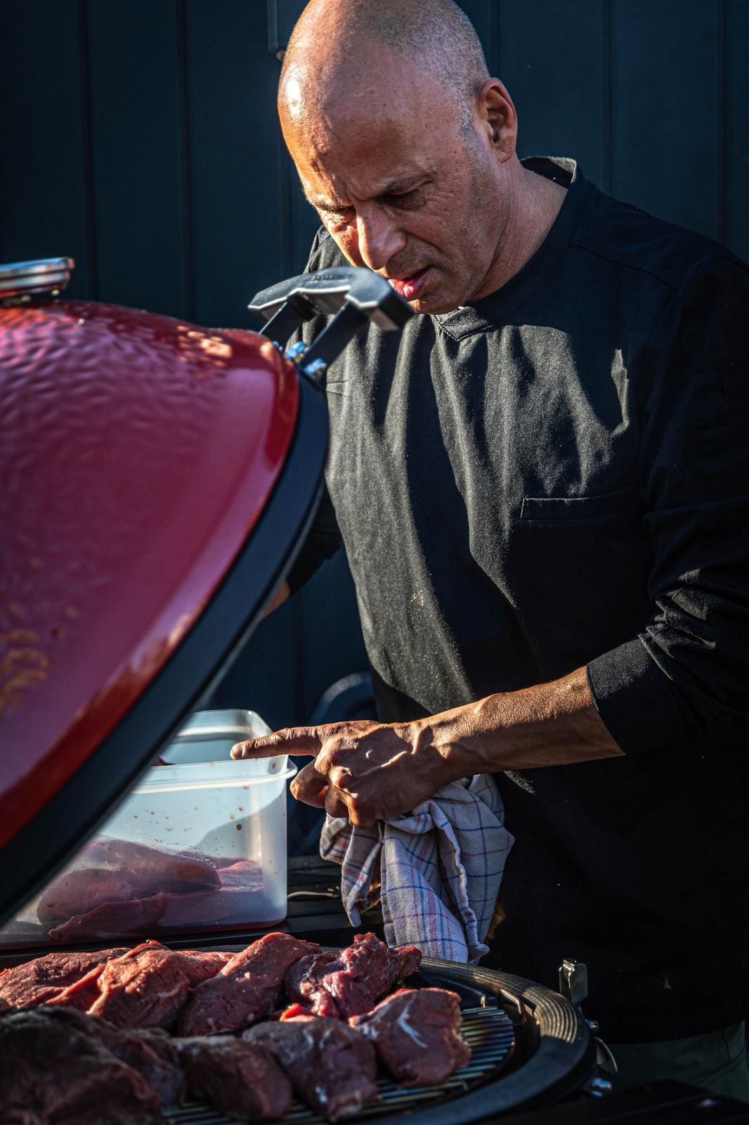 Een man grilt steaks op een rode barbecue en houdt een handdoek naast een open deksel.
