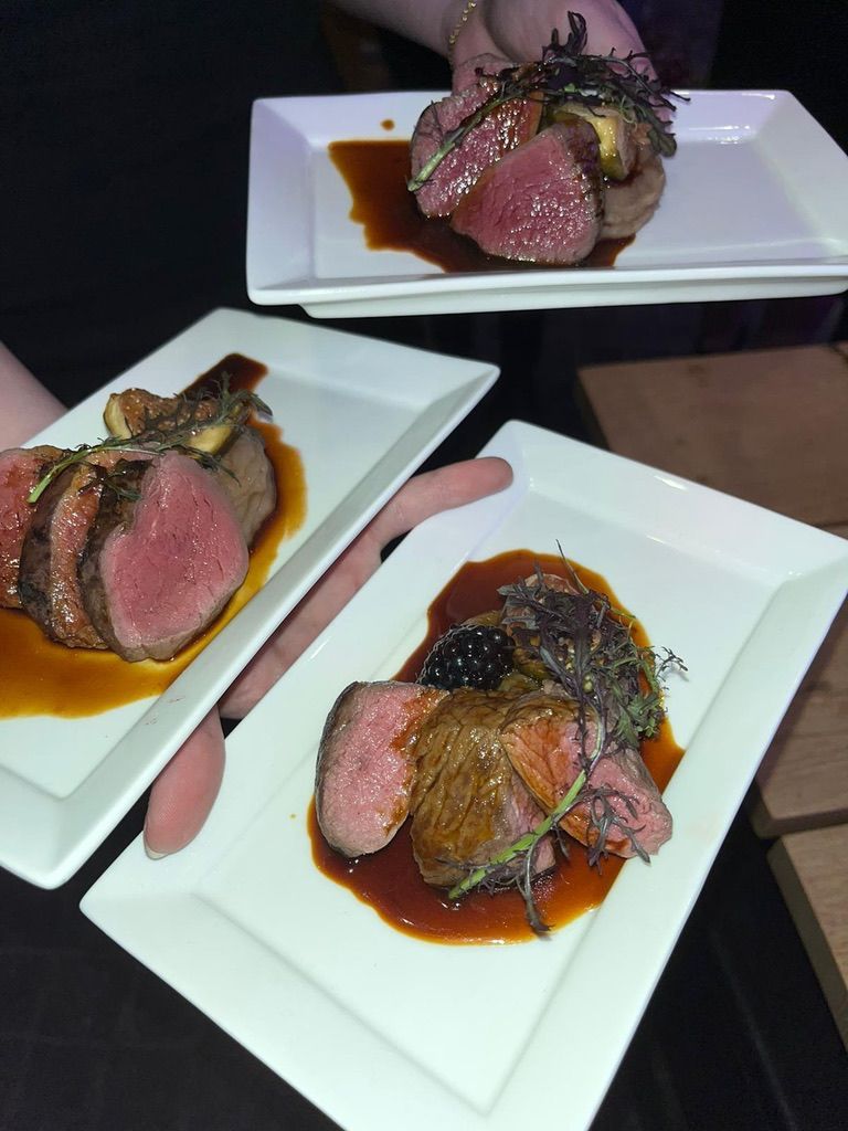 Plakjes aangebraden biefstuk met saus en kruiden, geserveerd op witte, rechthoekige borden in een schemerig restaurant.