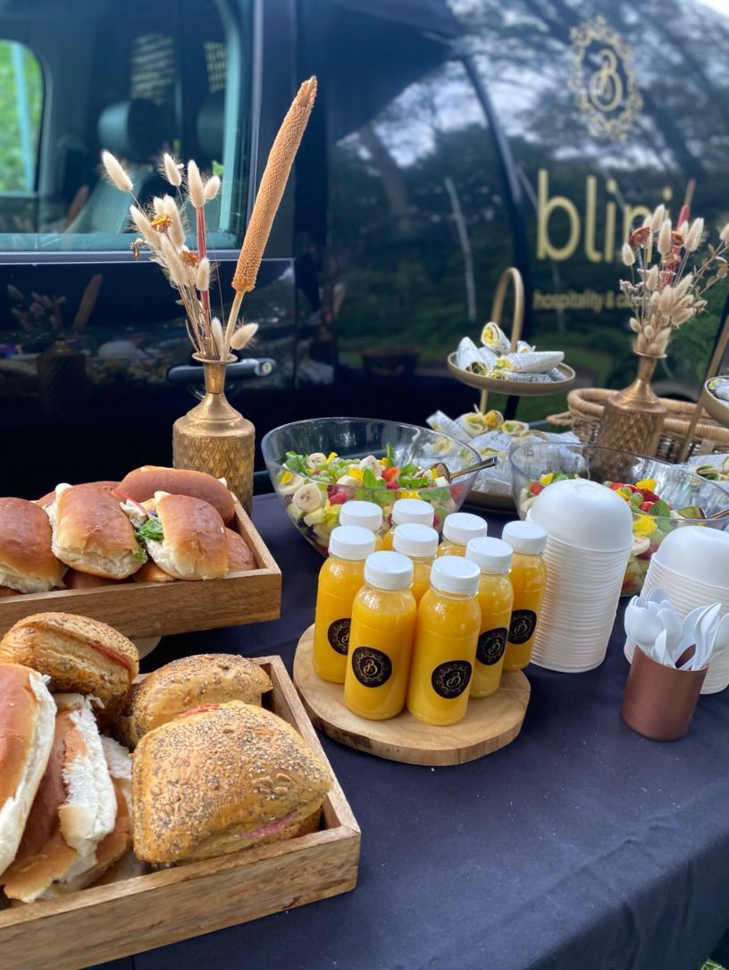 Ontbijtbuffet met gebak, brood, sinaasappelsap in flesjes en koffie op een tafel buiten.