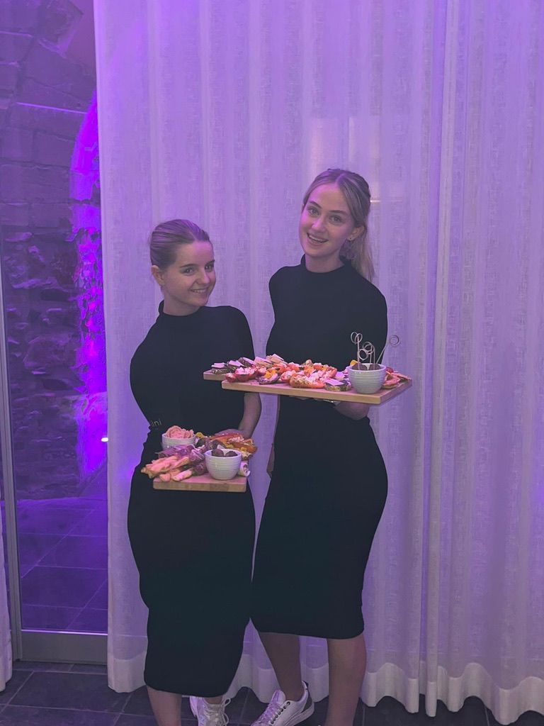 Twee vrouwen in zwarte jurken houden versierde dienbladen vast in een paars verlichte kamer met witte gordijnen.