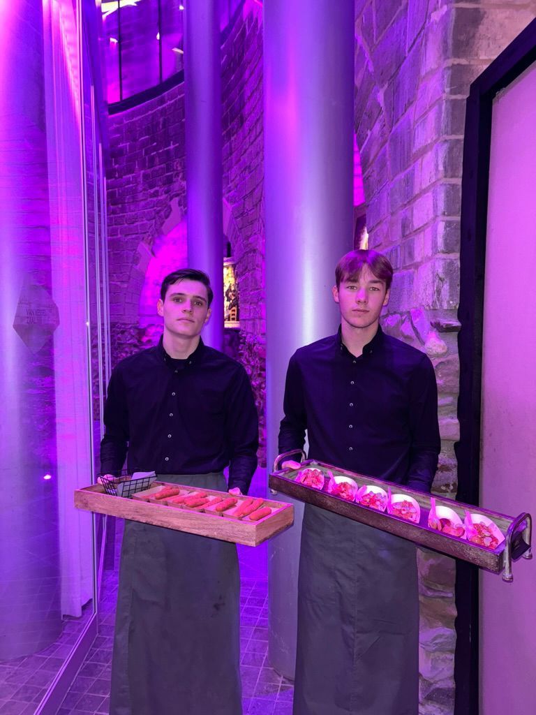 Twee obers met dienbladen vol eten onder paarse verlichting in een smalle stenen gang.