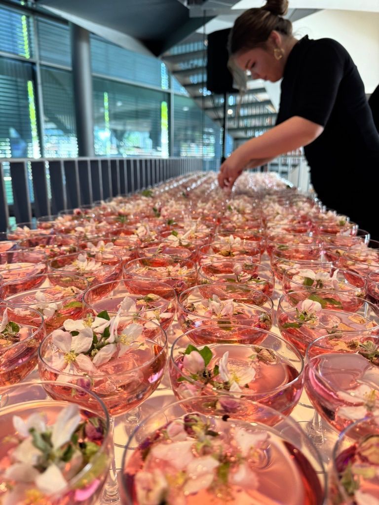 Rijen roze cocktailglazen op een buffettafel, die door iemand worden gerangschikt in een lichte, overdekte ruimte.