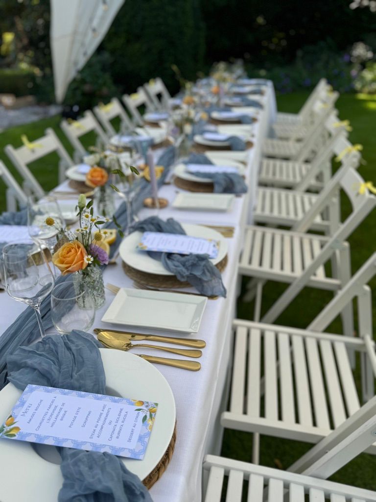 Lange eettafel voor buiten, compleet met witte stoelen, donkerblauwe servetten, gouden bestek en bloemaccenten.