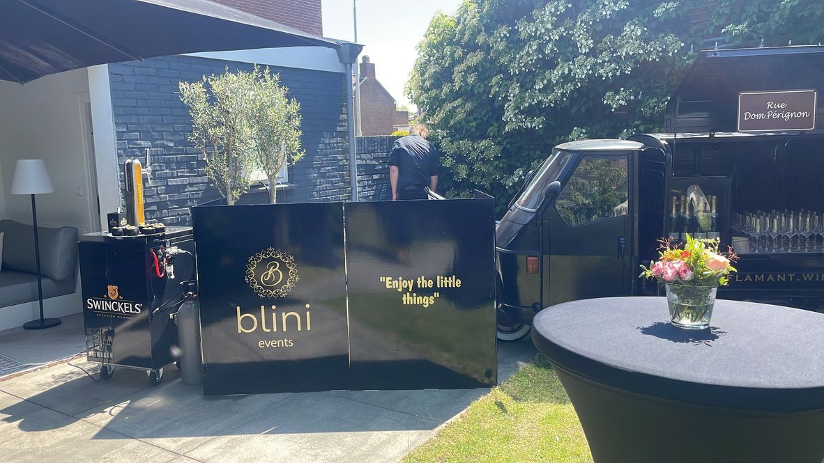 Een buitenpresentatie met zwarte bewegwijzering, een bloemstuk en een ronde tafel op een zonnig terras.