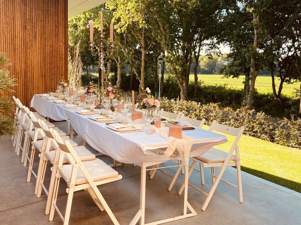 Lange eettafel voor buiten met witte stoelen onder bomen naast een houten muur.