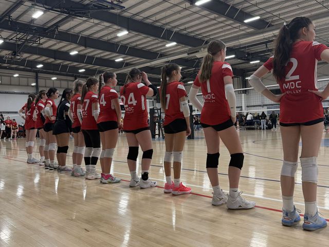 Volleyball players in red jerseys lined up in an indoor gym, waiting on the court.