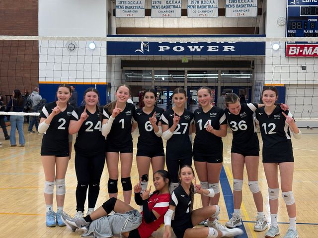 Women’s volleyball team posing in black uniforms in a gym, with two players kneeling in front