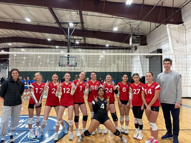 Volleyball team posing on an indoor court, with players in red uniforms and one in black crouching front center.