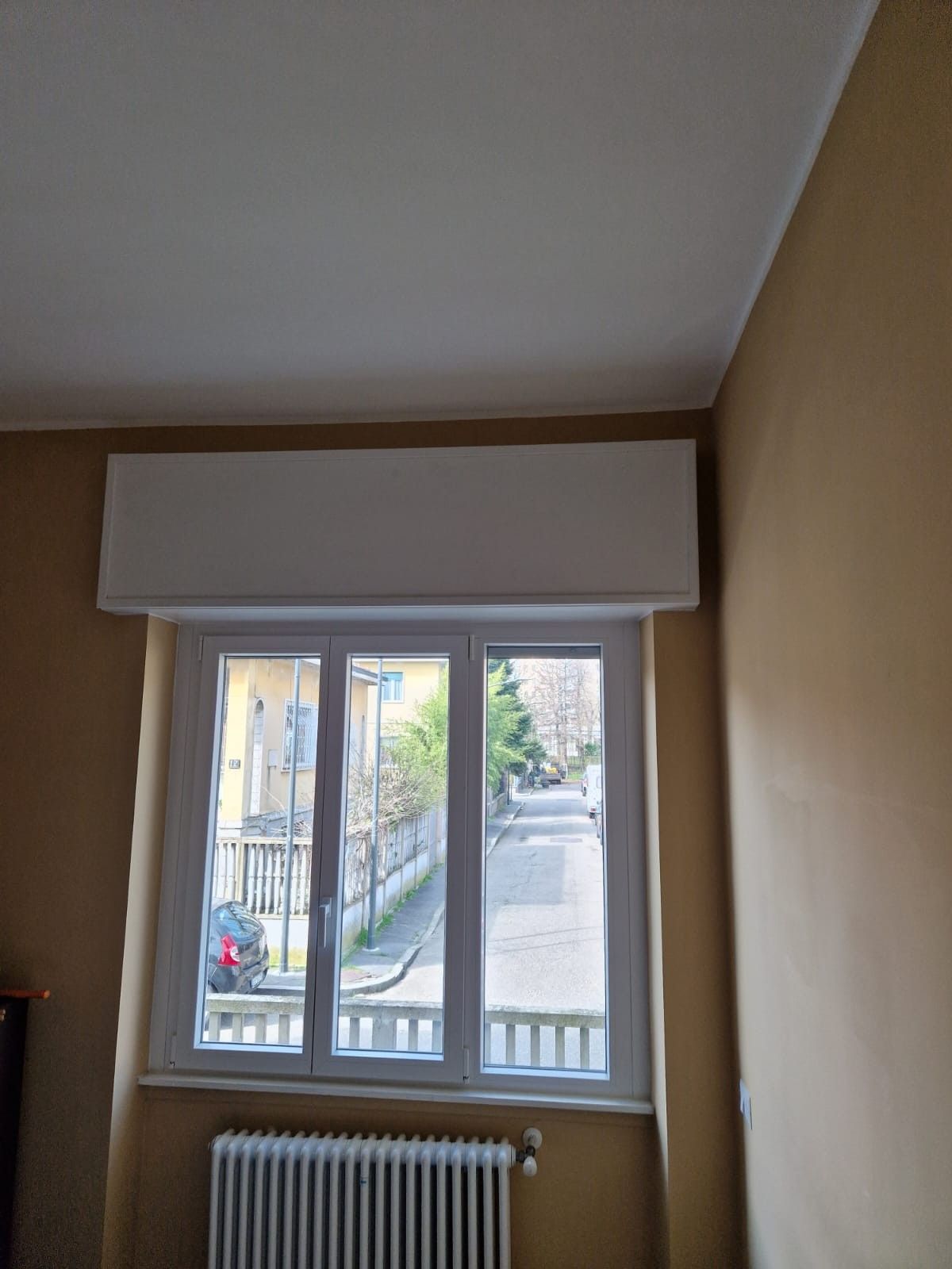 Vista interna di una finestra con tenda bianca, termosifone sottostante, pareti beige e scena di strada all'esterno.