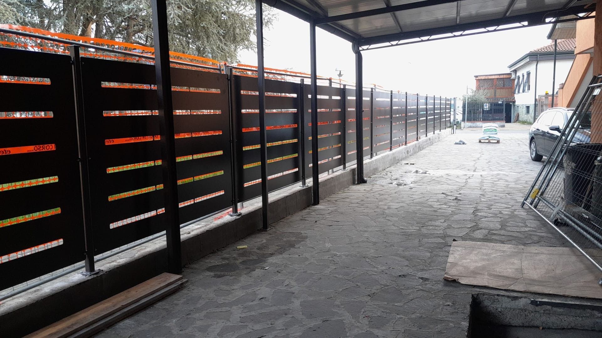 Stretto vicolo industriale con scaffalature metalliche nere sulla sinistra e un terreno innevato sotto una tettoia.