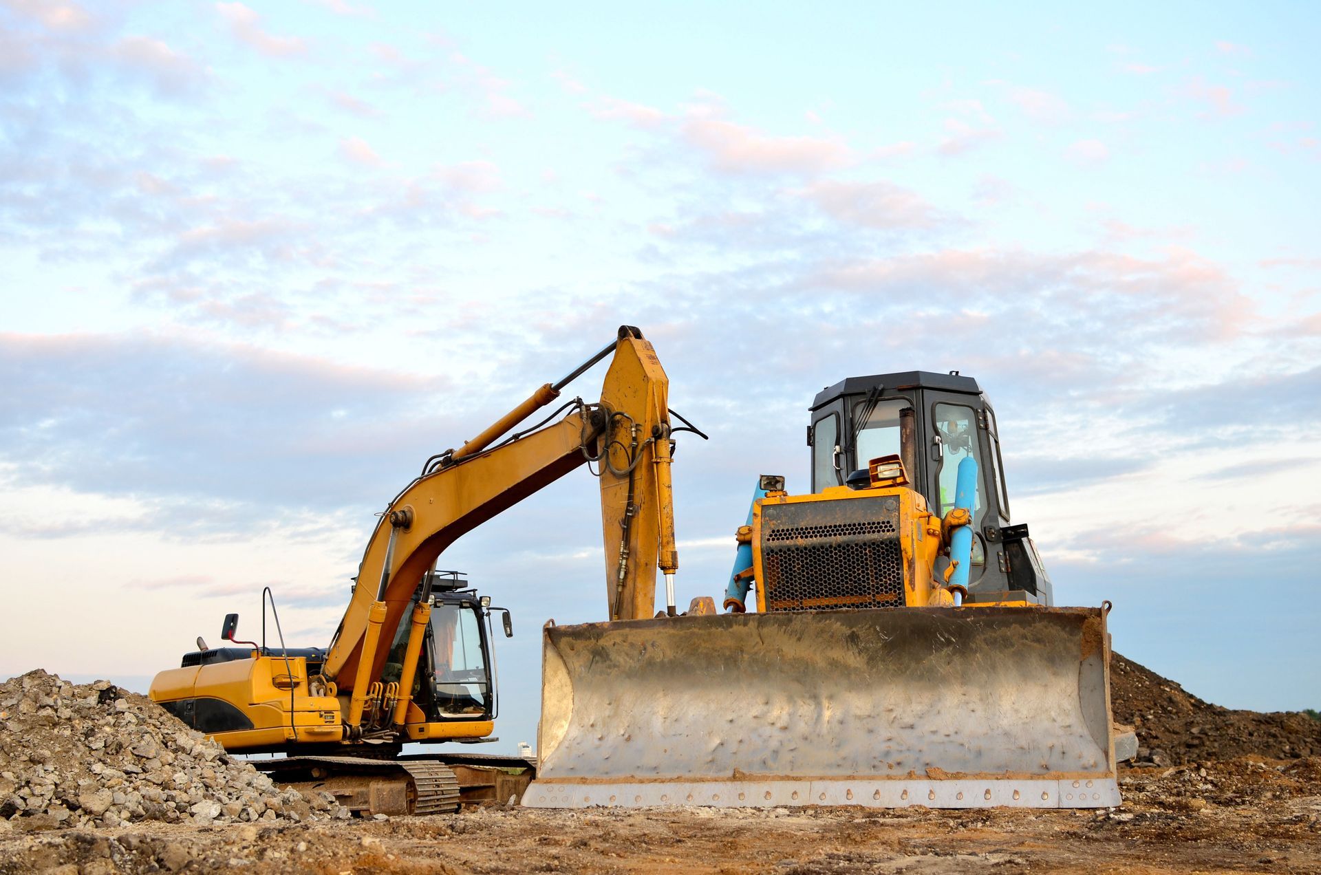Two yellow excavators on a dirt site, one with a large front blade, under a cloudy sky