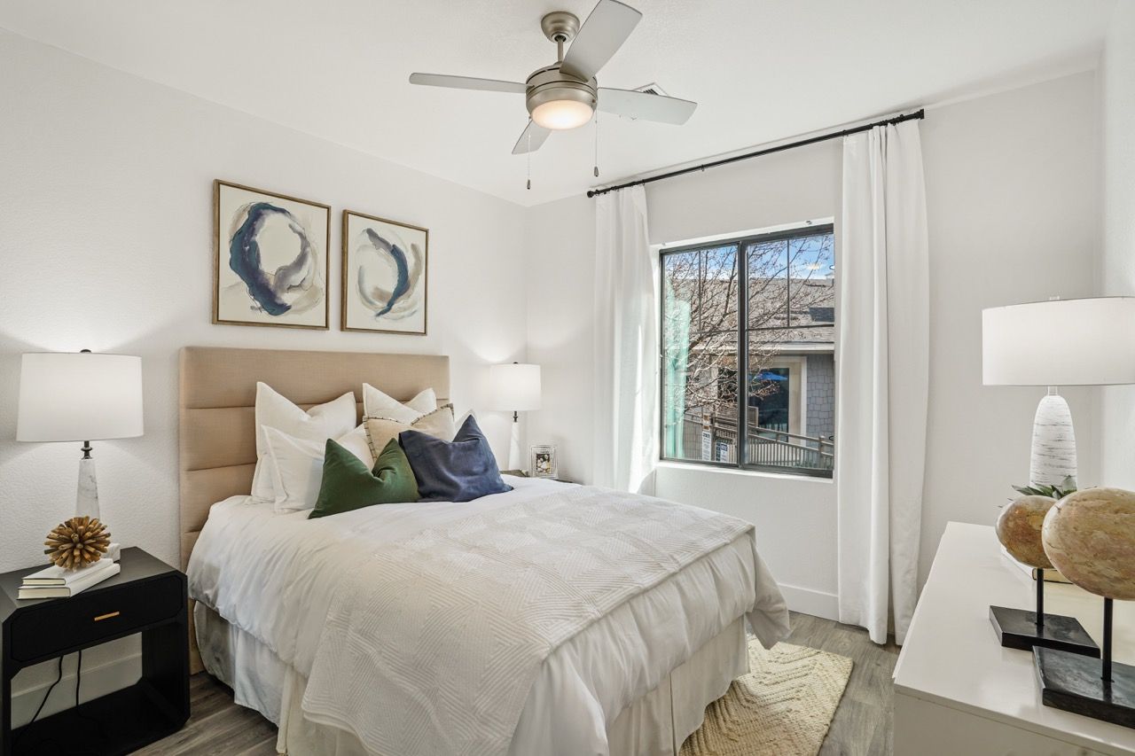 Bedroom in an apartment with a bed, nightstands, lamps, and a window with white curtains.