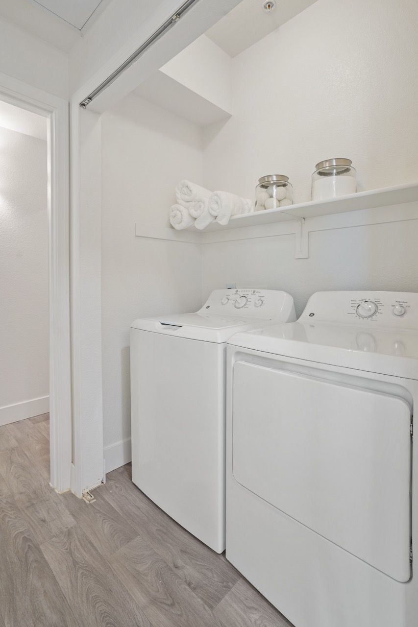 White washer and dryer in a compact apartment laundry room with a shelf of rolled towels and jars.