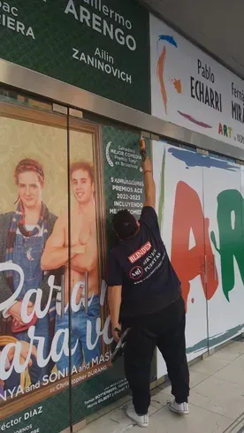 Persona limpiando el escaparate de una tienda con carteles y letreros en español.