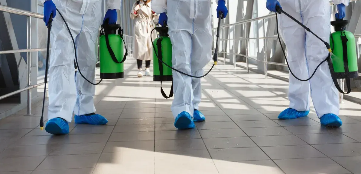 Three people in white hazmat suits, blue booties, and gloves spray a hallway. A person in a tan dress walks behind them.