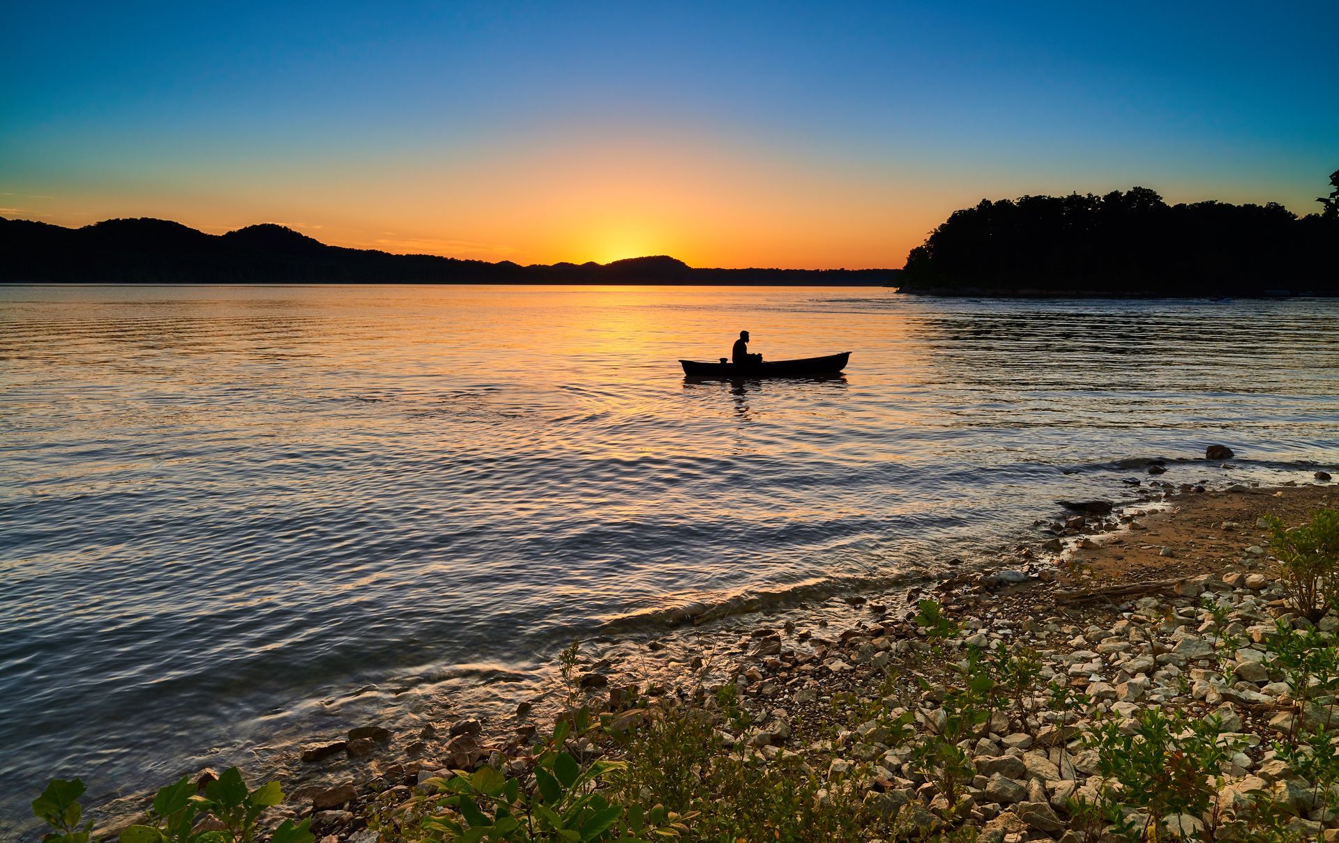 Sunset over a lake; a person paddles a boat toward the horizon.