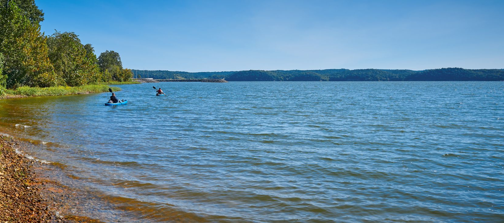 Two people paddle on a lake near a rocky shore with trees and a forest on the distant horizon under a blue sky.