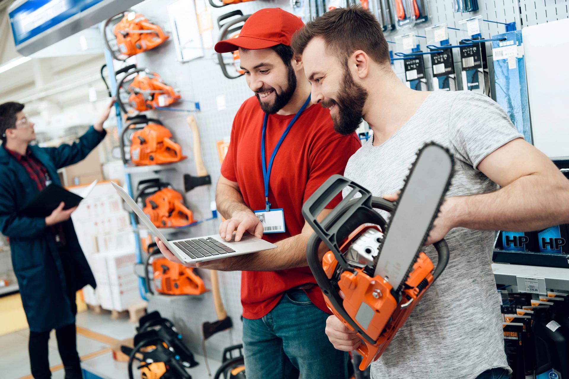 Two men looking at a chainsaw in a hardware store. One holds a laptop.