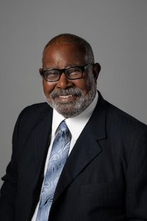 Studio portrait of a smiling man in a dark suit, white shirt, and patterned blue tie on a gray background