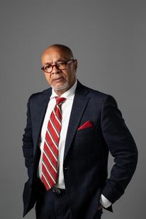 Portrait of a man in a dark suit with a red striped tie and pocket square, standing against a gray background