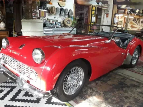 Red vintage sports car in a cluttered garage, viewed from the front-left.