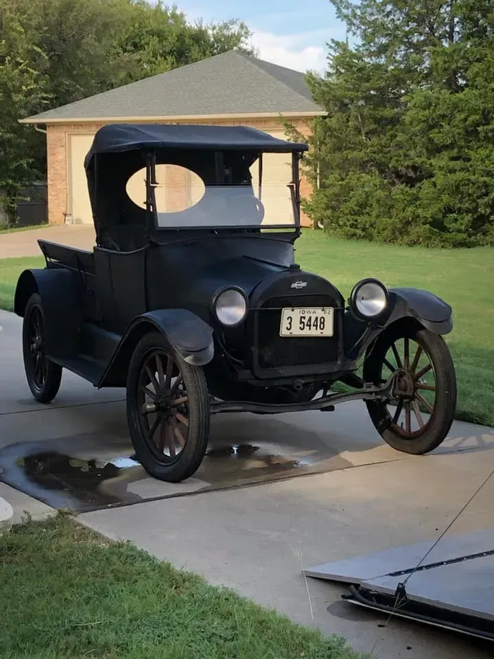 Black vintage pickup truck parked in a driveway beside a house, with trees in the background