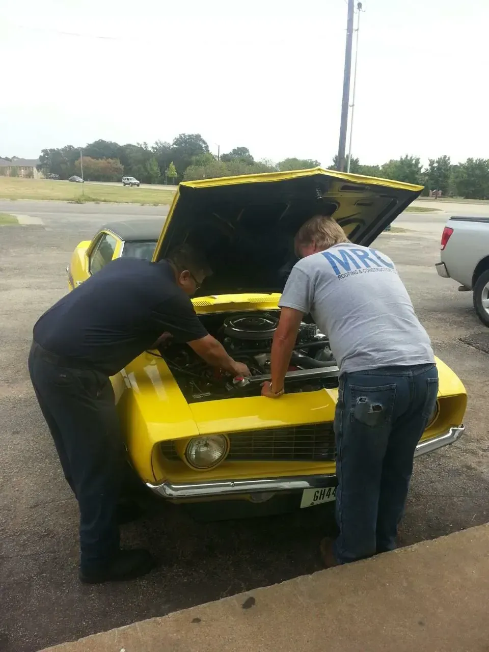 Two people leaning over a yellow car with the hood open, inspecting the engine in a parking lot.
