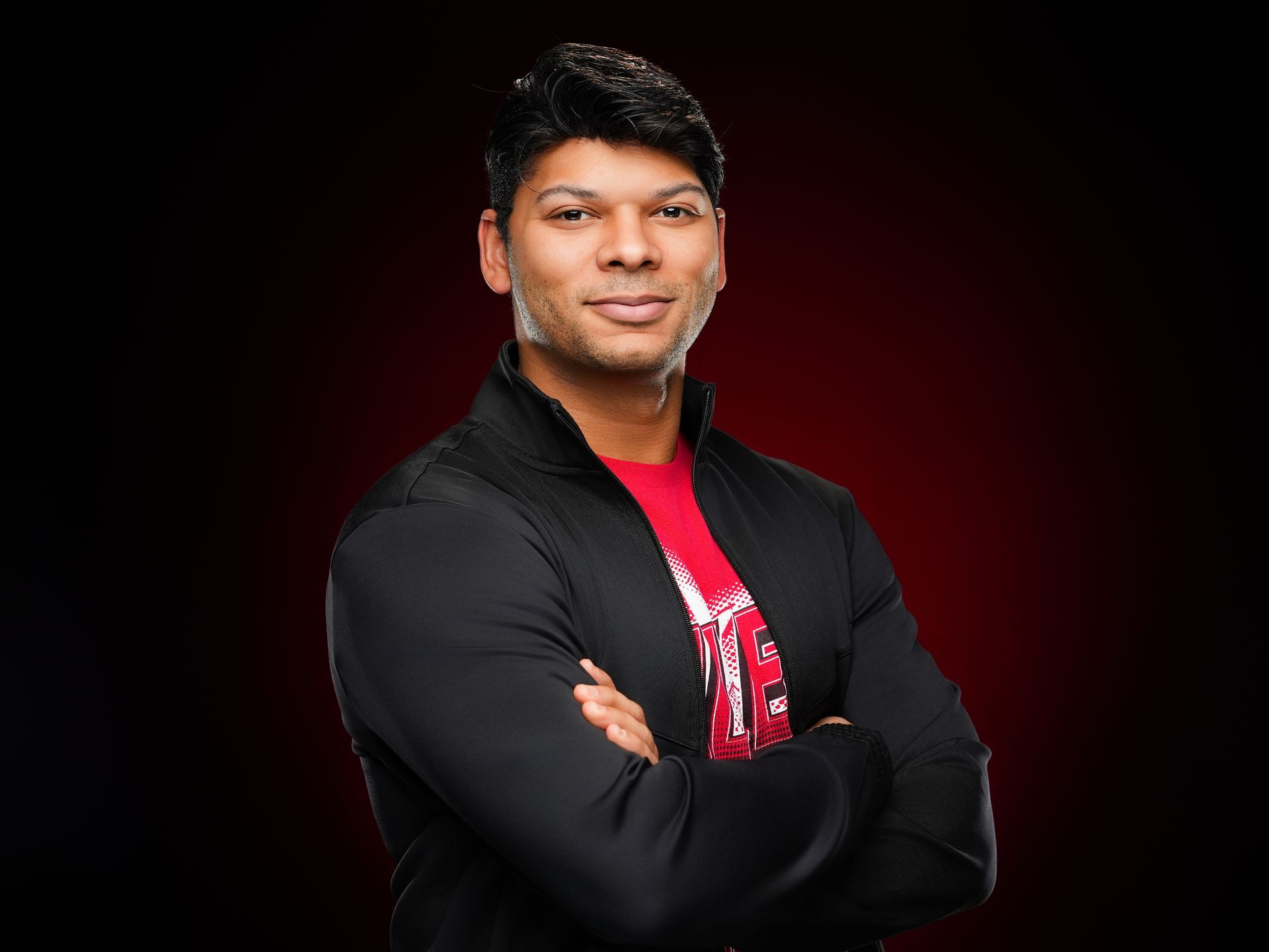 A person with dark hair wears a black zip-up jacket over a red graphic t-shirt, arms crossed, against a dark red background.
