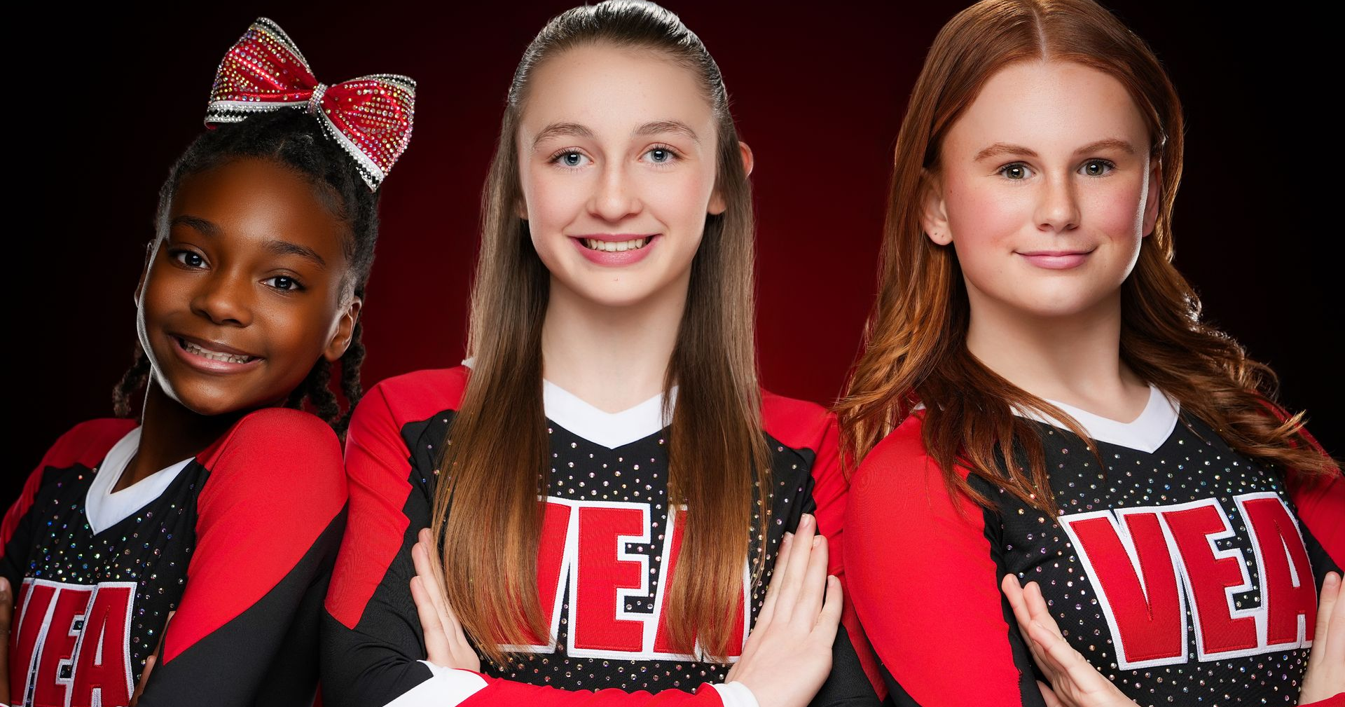 Three cheerleaders in red and black uniforms pose against a dark red background, smiling with their arms crossed.