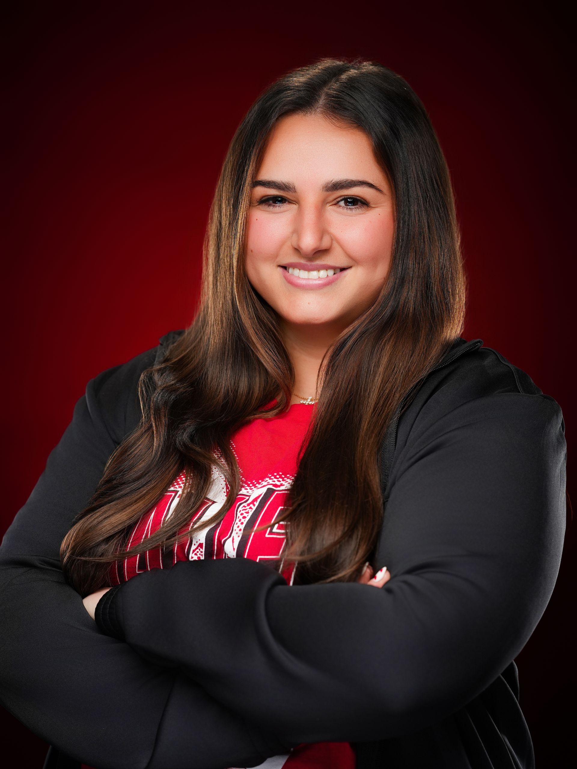 Smiling person with long, dark, wavy hair, wearing a black jacket over a red shirt, arms crossed against a red background.