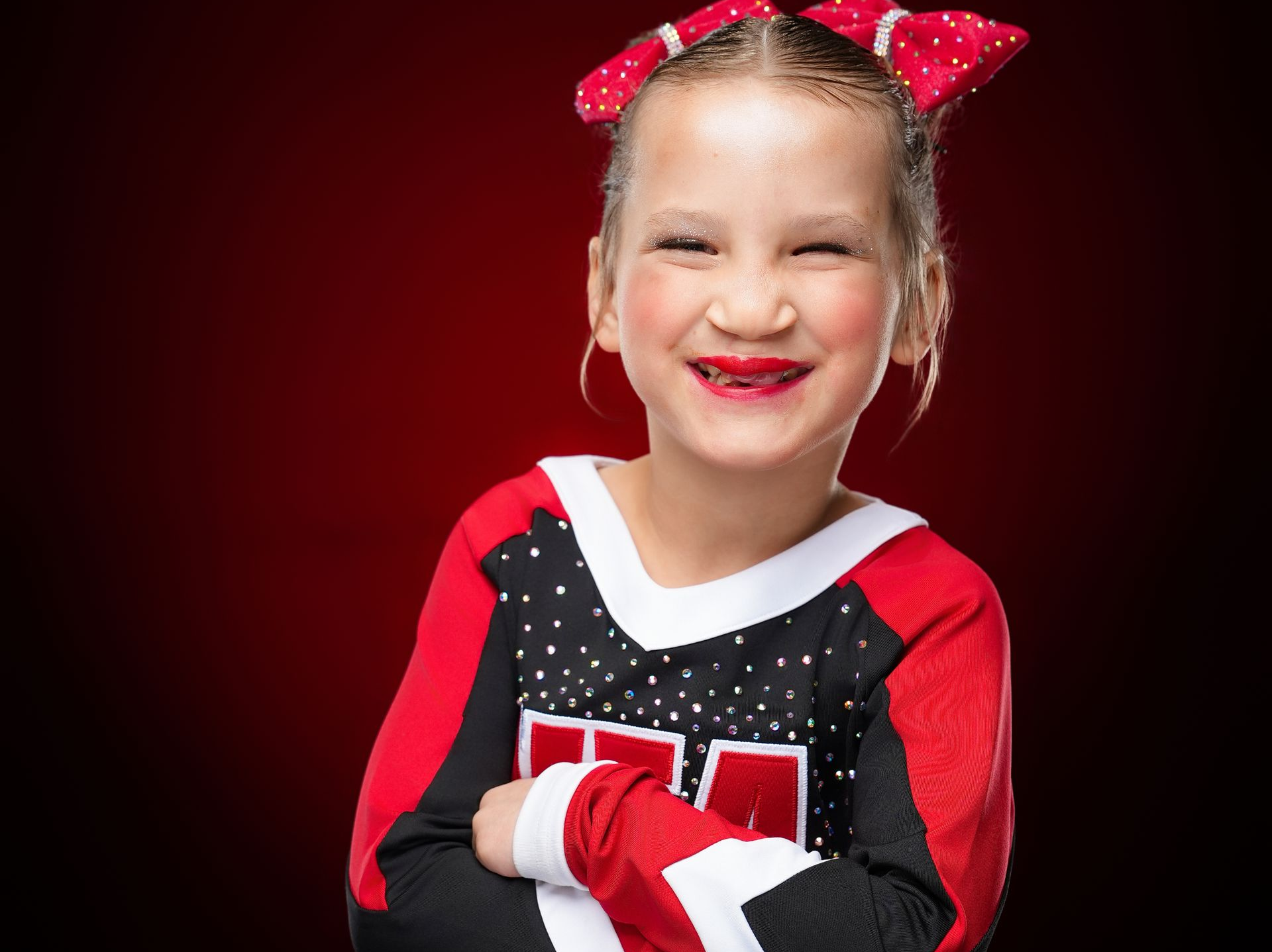 A smiling person with a red bow in their hair, wearing a red and black long-sleeved cheerleading uniform, against red.