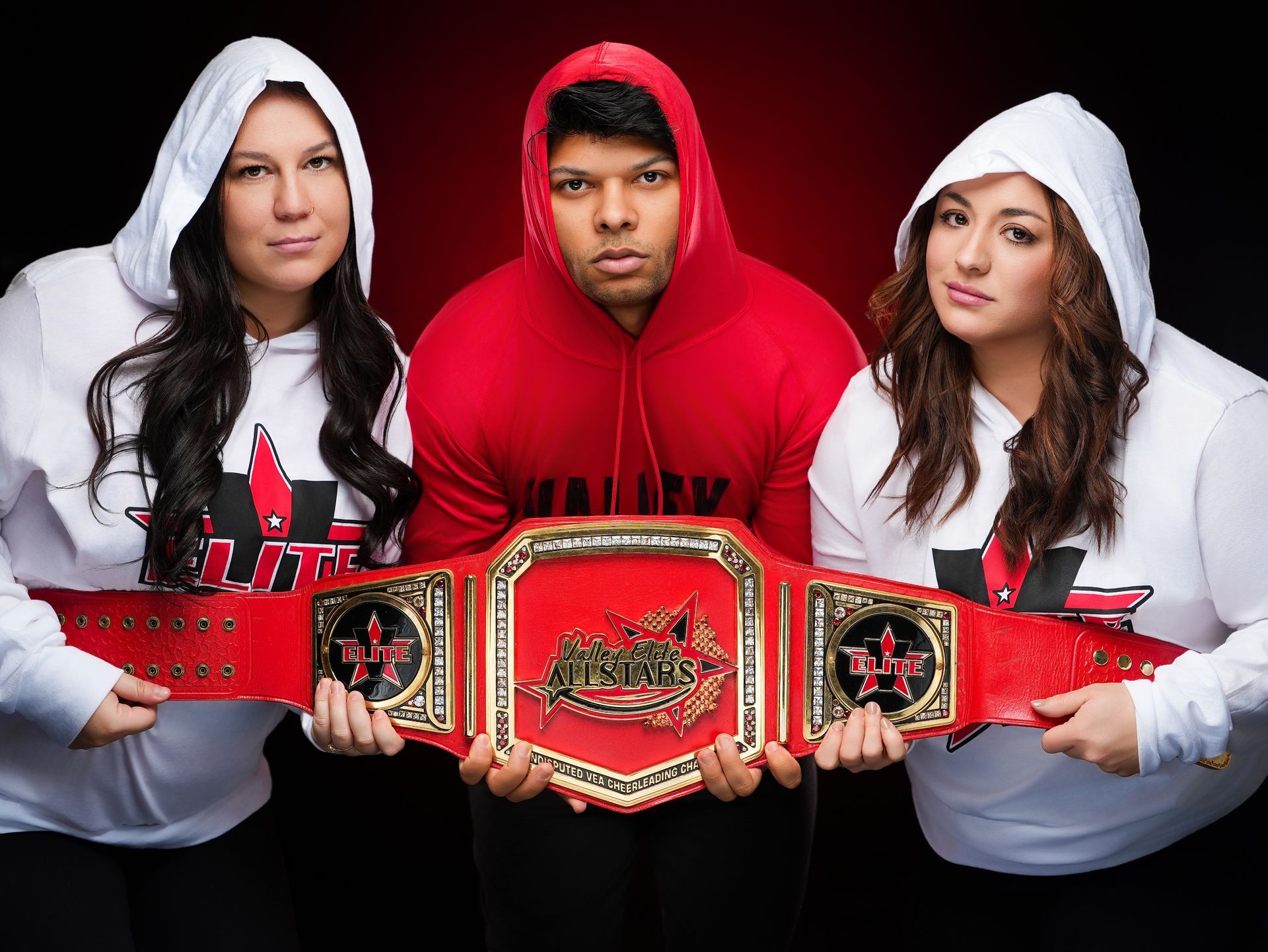 Three people wearing hooded sweatshirts pose together, holding a large championship belt with a red strap.