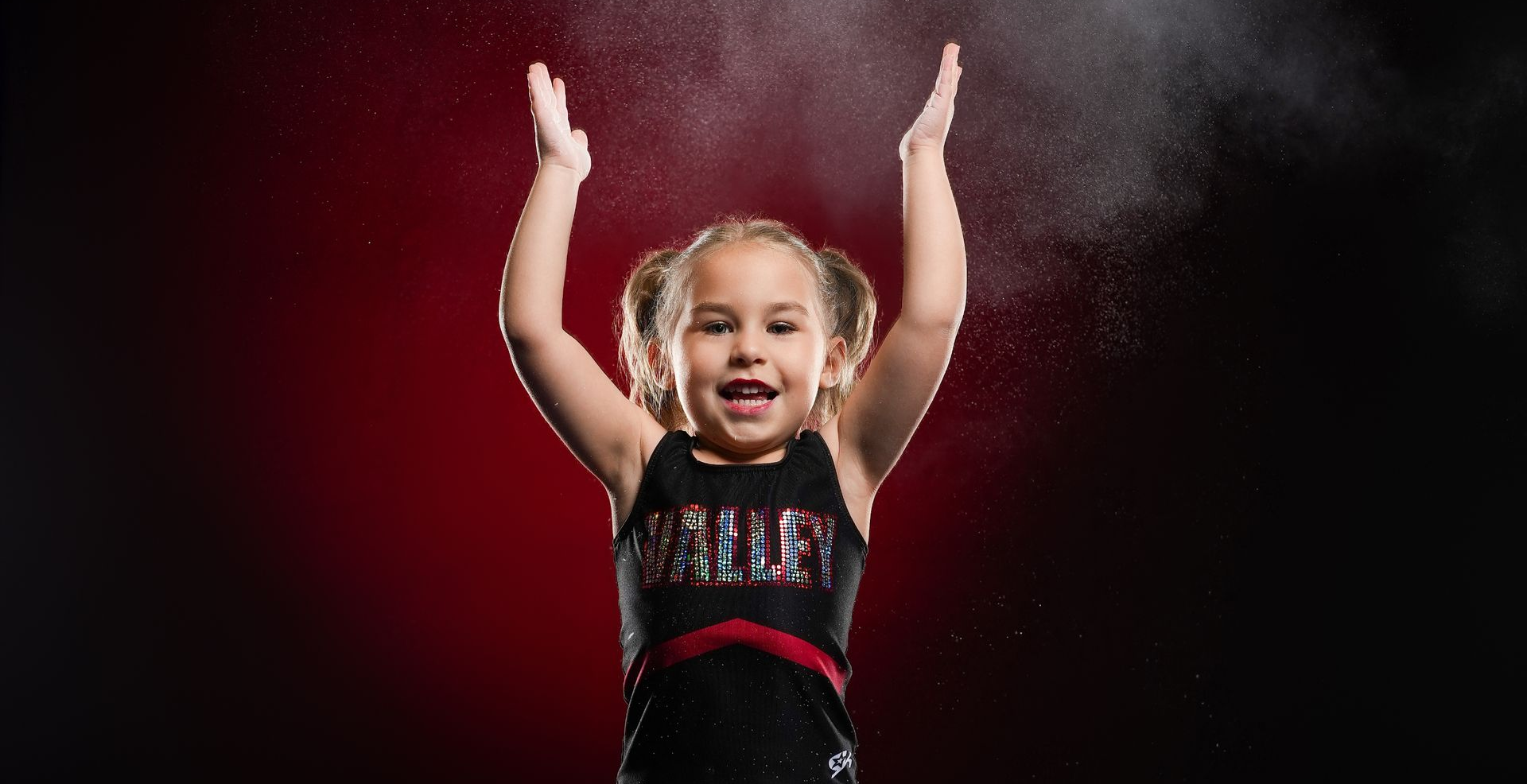 A young gymnast in a black and red leotard claps her hands, creating a spray of white chalk against a dark red background.