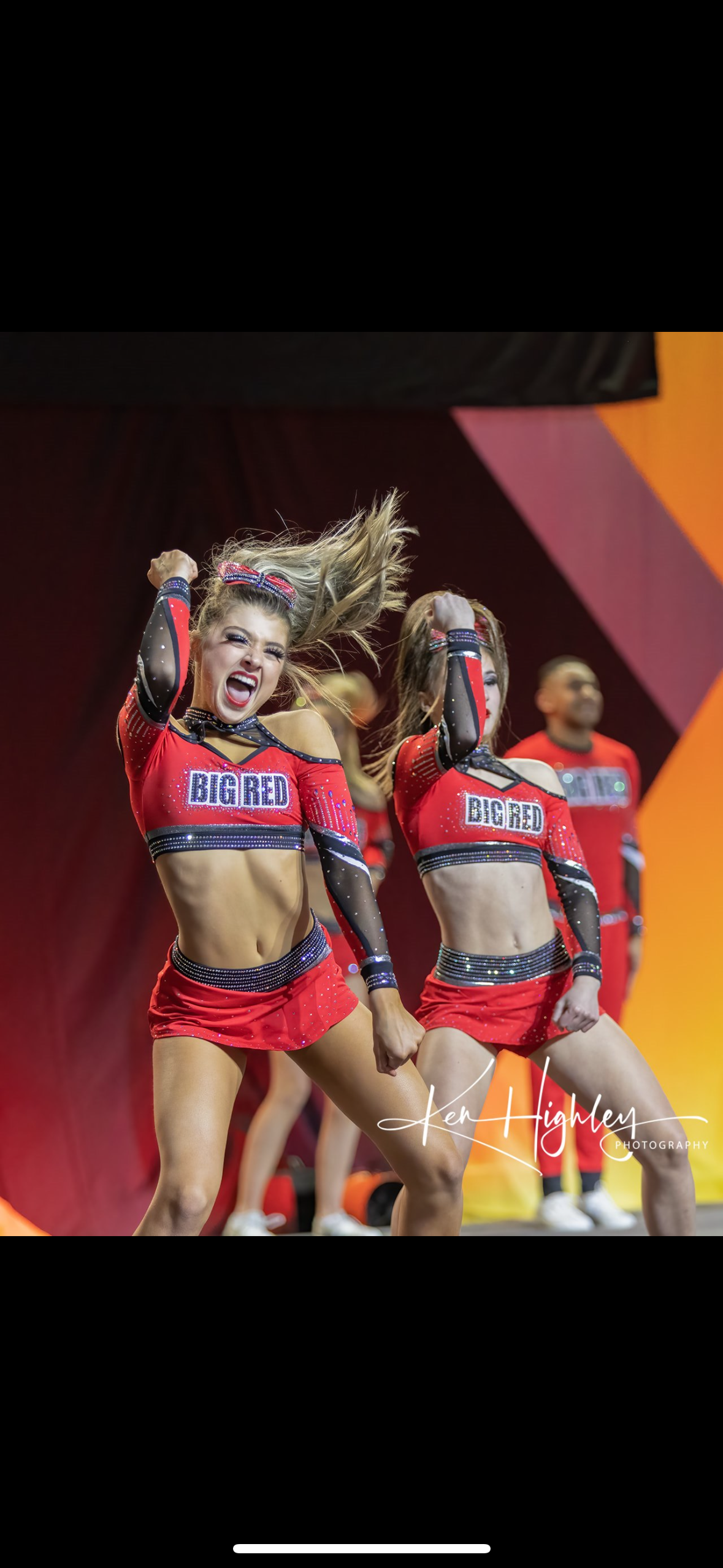 Two cheerleaders in matching red uniforms perform a routine on stage, expressing joy with their arms raised.