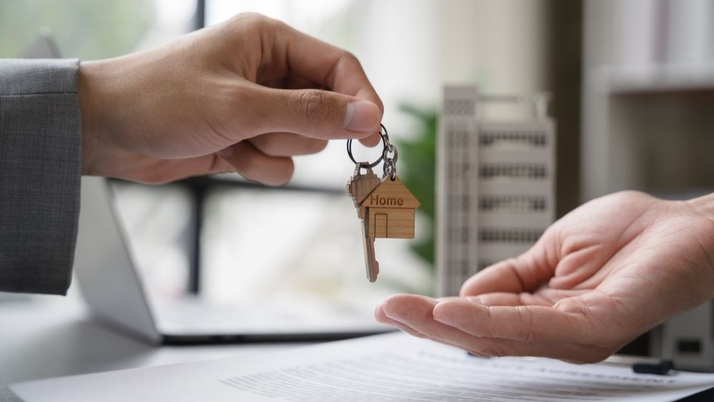 Hand handing over house keys. Blurred background includes a building model, laptop, and documents.